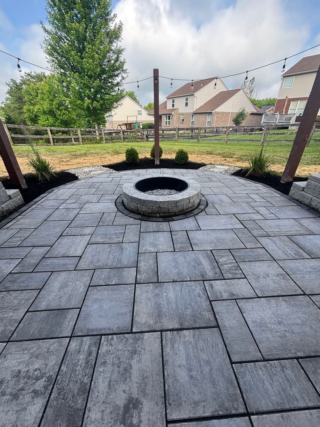 Patio with stone fire pit, surrounded by landscaping and houses in the background, under cloudy sky.