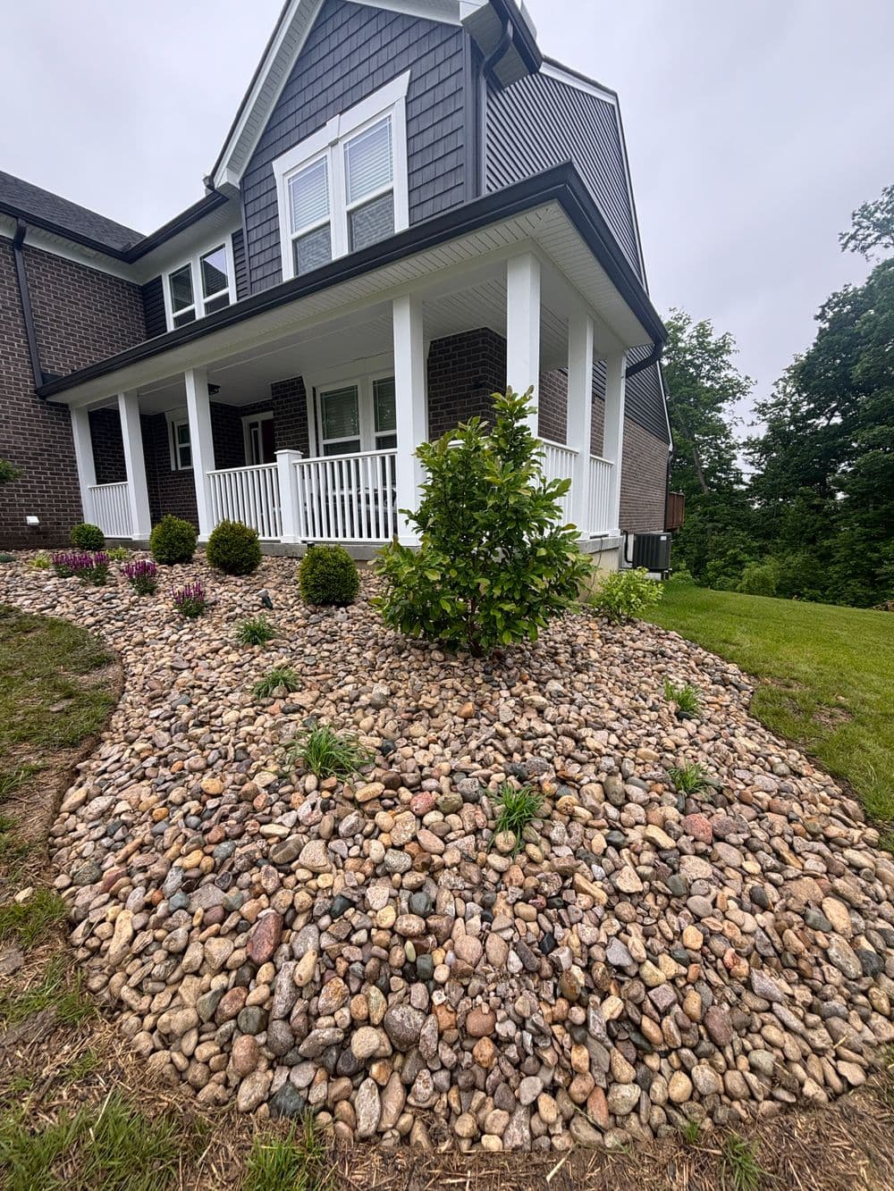 Landscaped stone garden with shrubs in front of a modern home. Green lawn and tree backdrop.