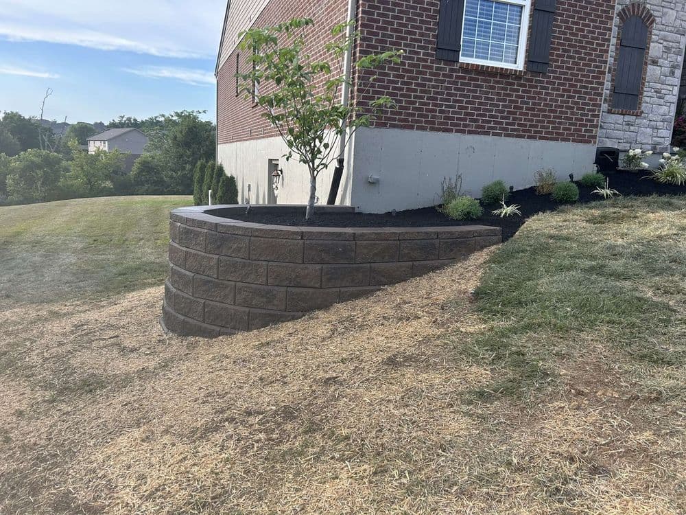 Landscape features a tiered stone wall with a young tree near a brick house and freshly laid sod.