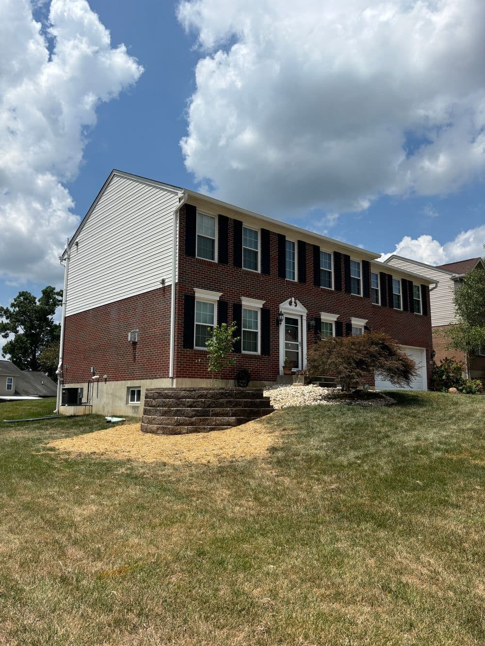 Two-story brick house with white siding, green lawn, and landscaping under a blue sky.