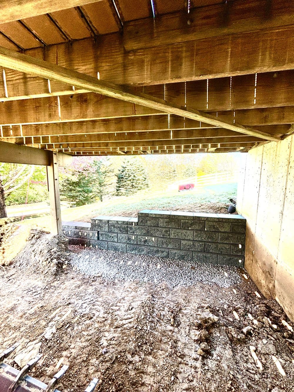 Under-construction space beneath a deck with grey retaining wall and gravel base.