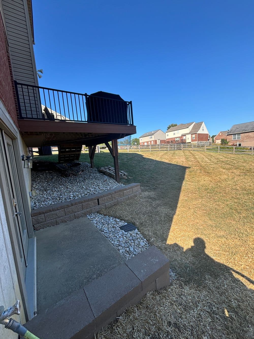 Backyard view featuring a deck, gravel area, and houses in the distance under a clear blue sky.