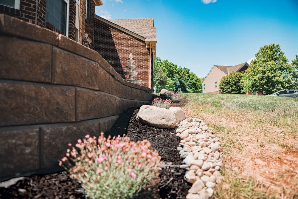 Flower garden with decorative rocks and landscaping wall near a brick house under clear blue sky.