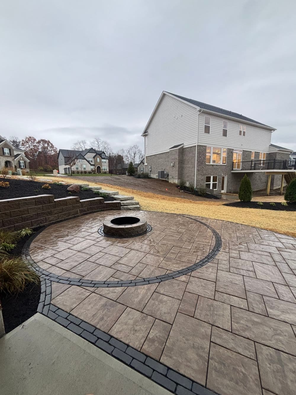 Patio with circular fire pit and textured pavers, surrounded by modern homes and overcast sky.